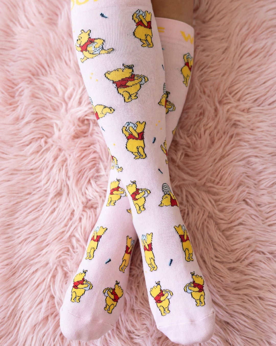 Crossed legs wearing pink Winnie the Pooh patterned socks on a fluffy pink rug