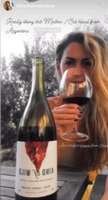 Person holding a wine glass with a bottle labeled 'Kind of Wild' on the table.
