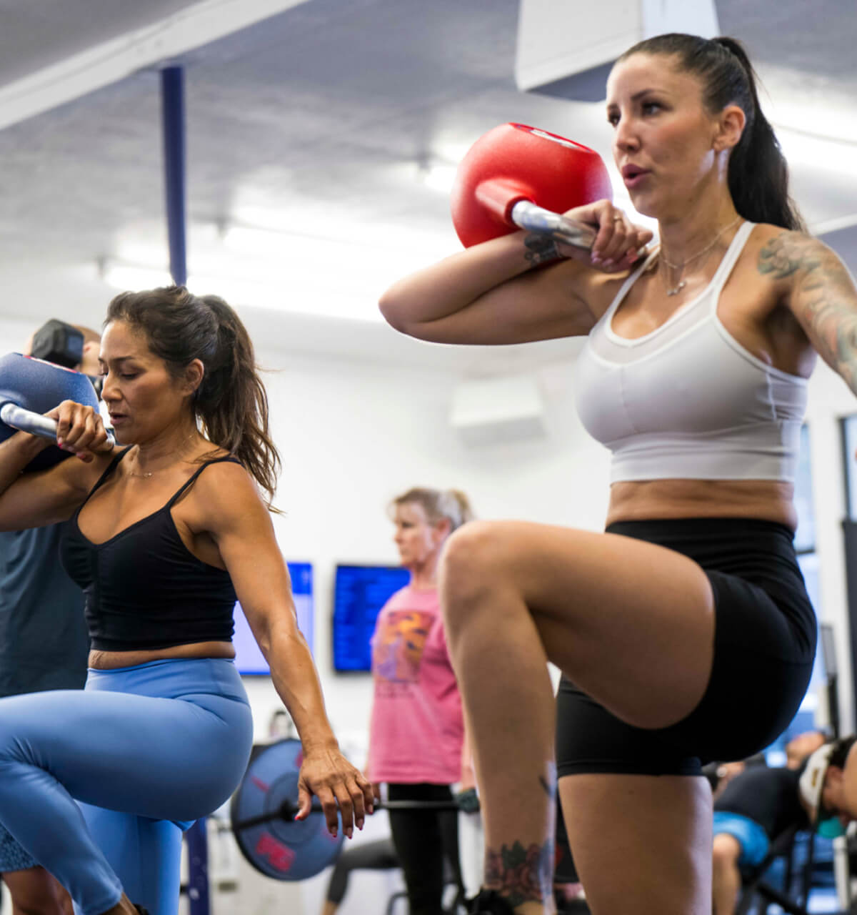 Two women exercising with barbells in a gym setting.