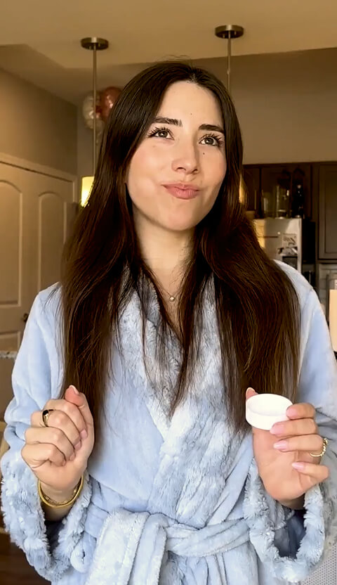 Woman in a blue robe holding a small white container in a kitchen.