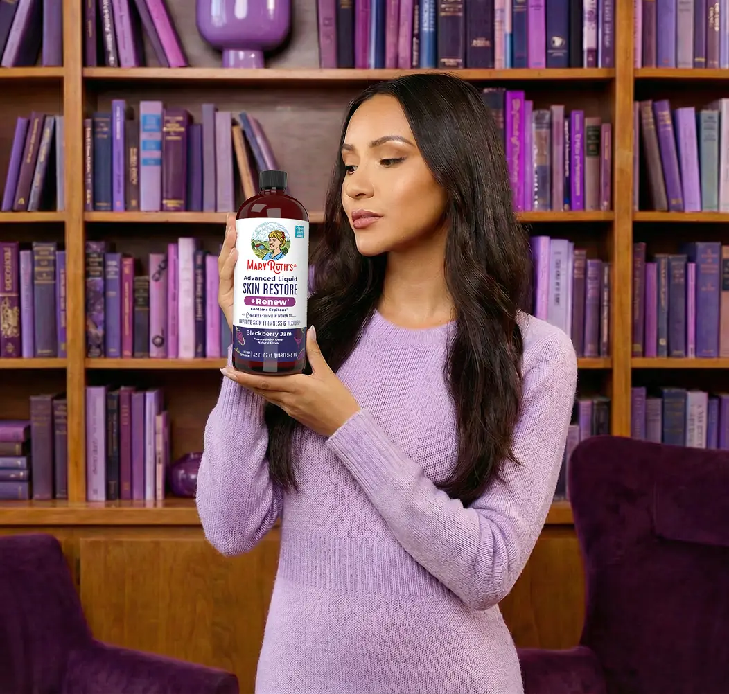 A woman in a purple sweater holds a bottle of a liquid supplement in front of a bookshelf.