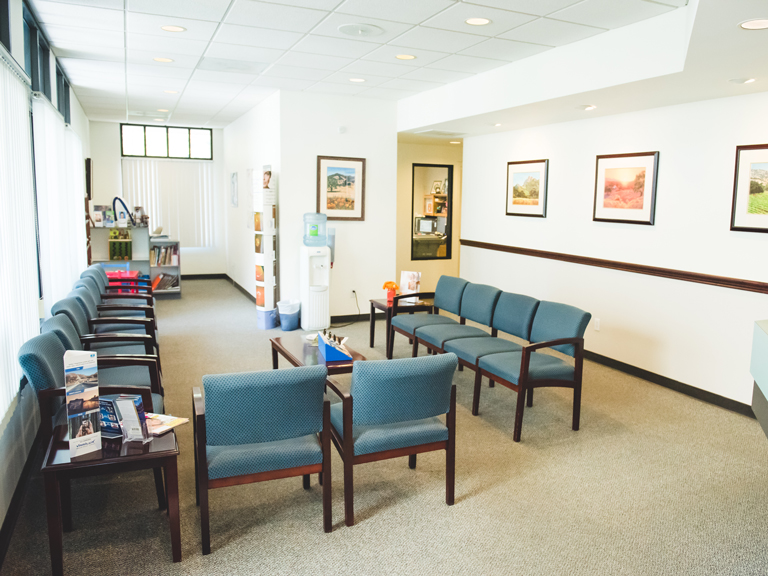 An empty, brightly lit office waiting room with rows of blue chairs and pictures on the walls.