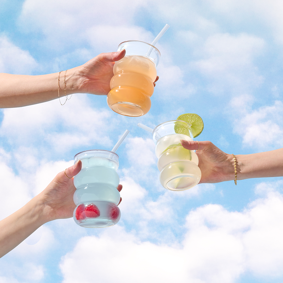 Three hands holding colorful drinks against a cloudy sky.
