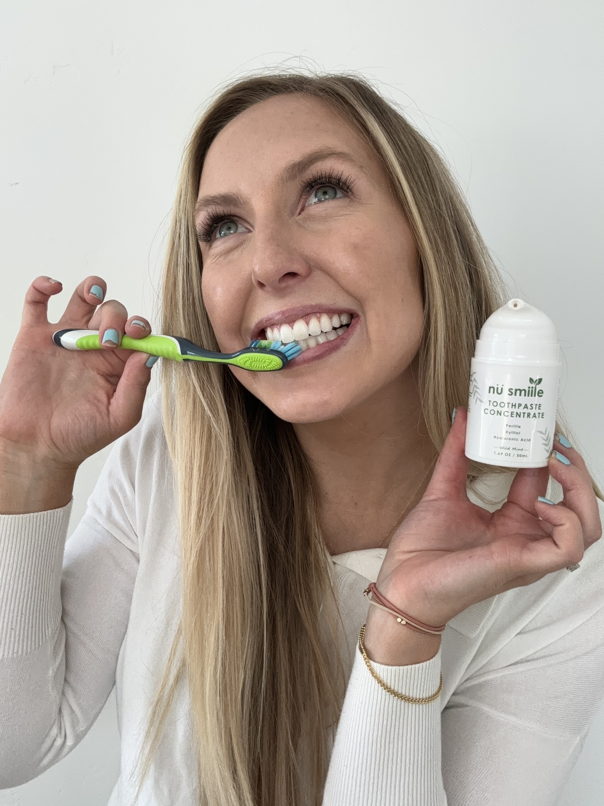 Person brushing teeth while holding toothpaste bottle, smiling.