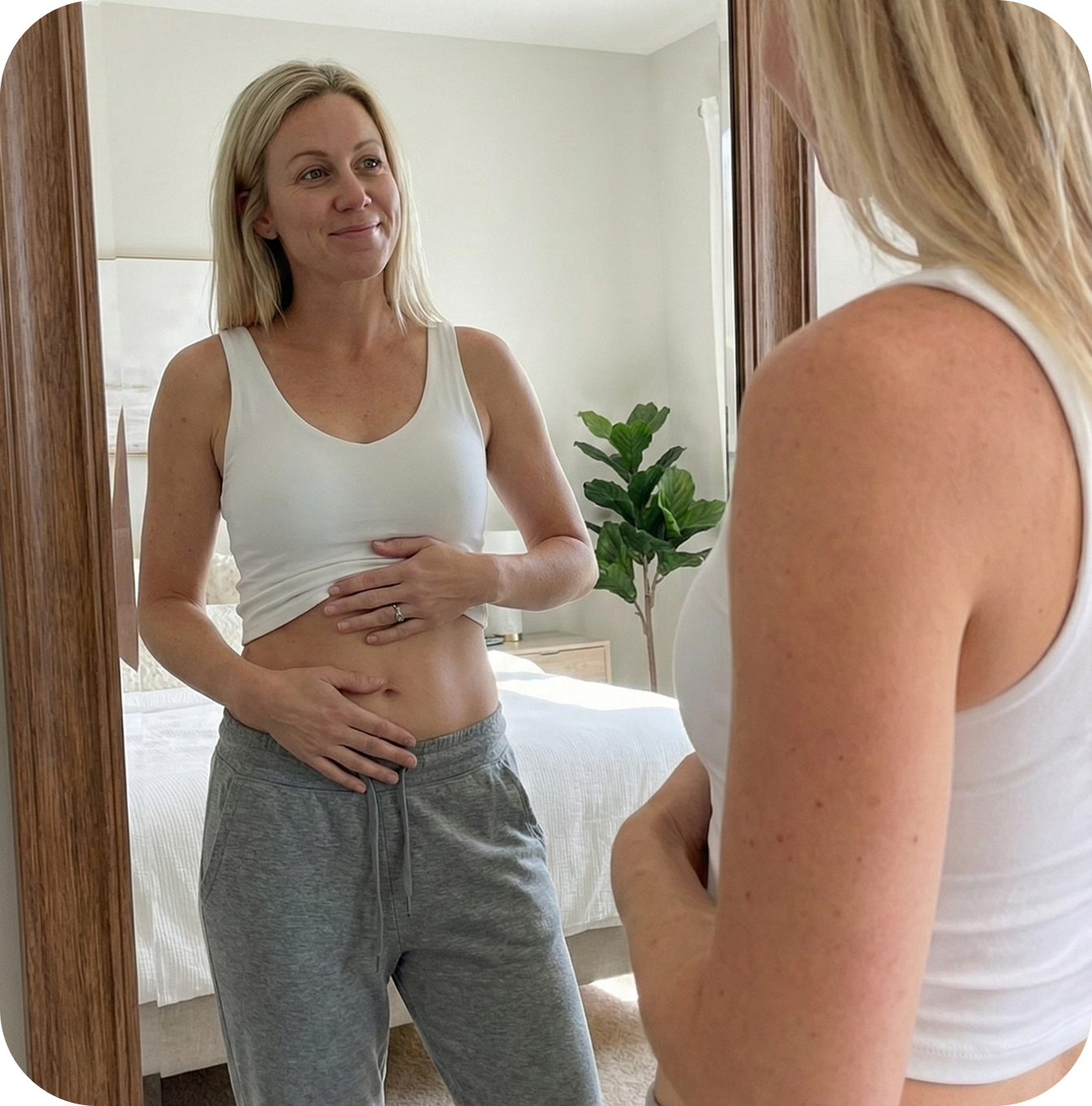 A woman in a white tank top and grey sweatpants looks at her reflection in a mirror.