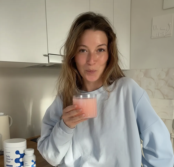 A woman in a light blue sweatshirt holds a glass of a pink drink in a kitchen.