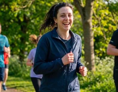 A woman in a blue jacket smiles while running in a park with other people.