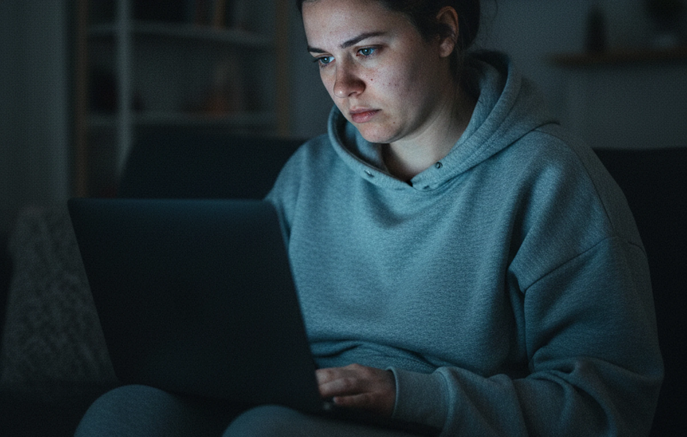Person in a hoodie focused on a laptop screen in a dimly lit room.