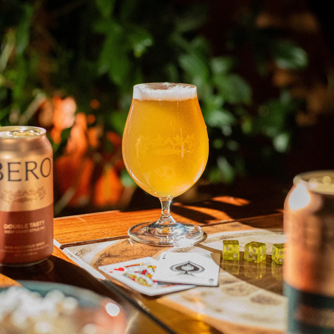 A glass of beer sits on a wooden table next to cans, playing cards, and dice.