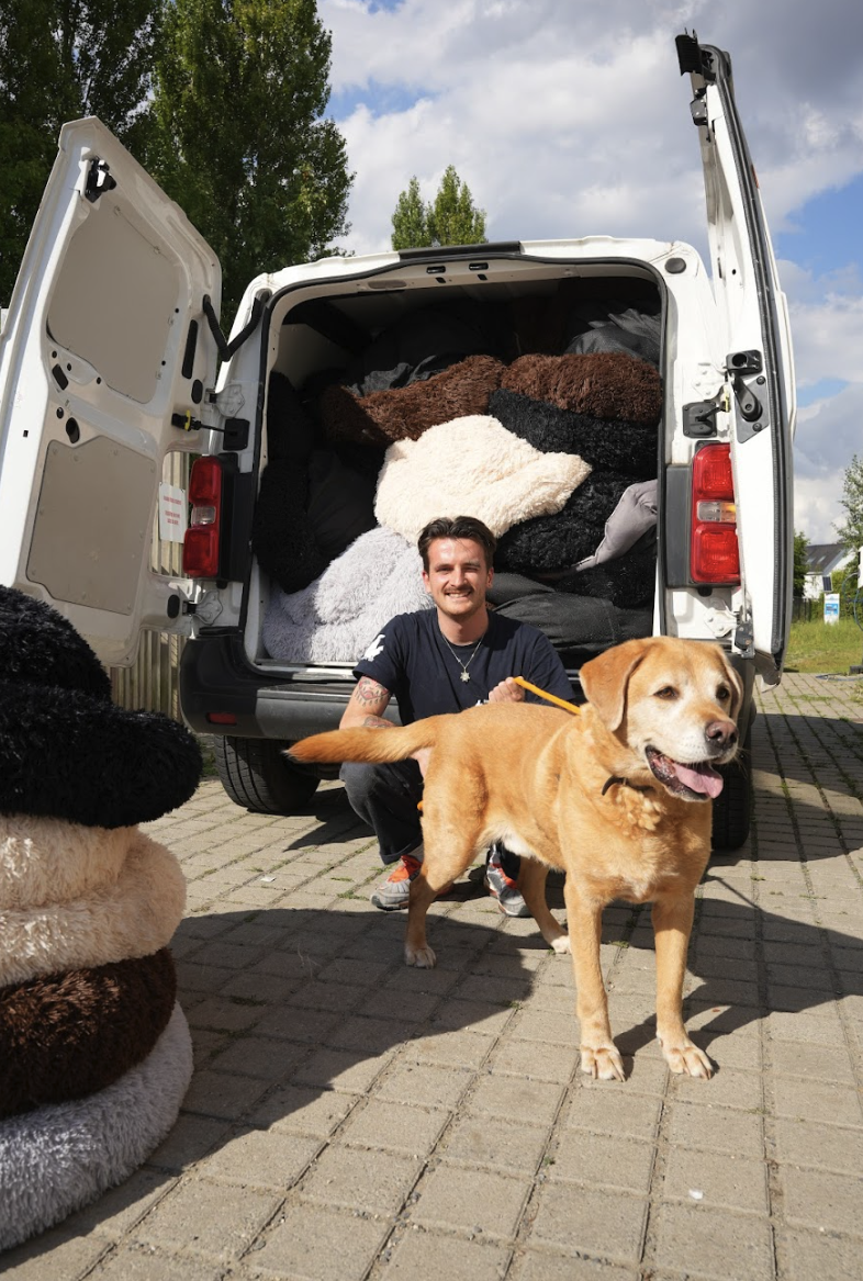A smiling man and a dog stand in front of a van filled with fluffy pet beds.