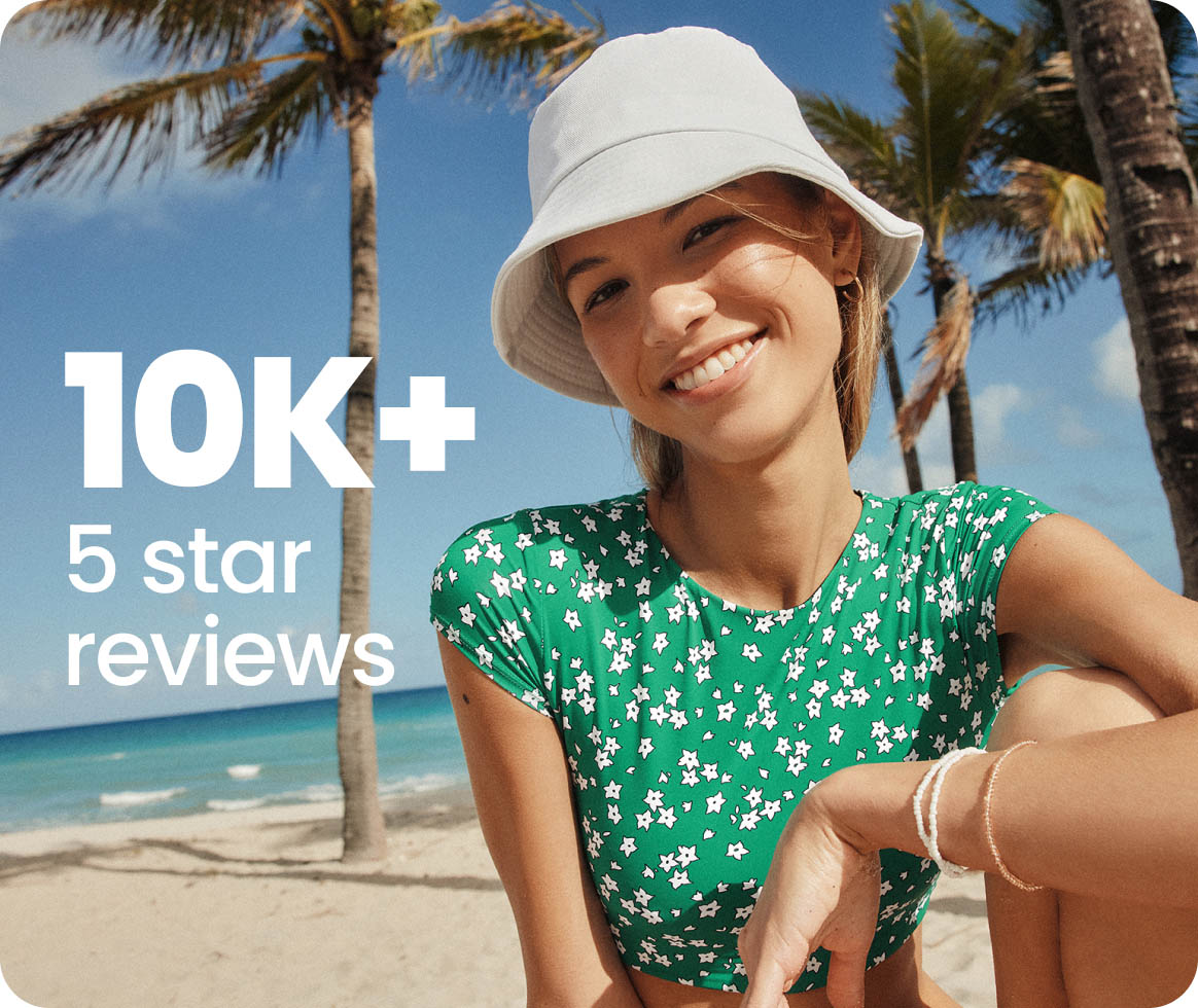 Smiling person on a beach with palm trees, wearing a hat and green floral top.