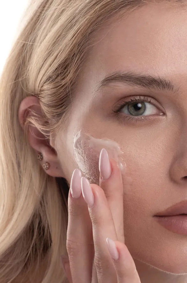 A close-up of a blonde woman with green eyes applying a clear gel to her cheek.
