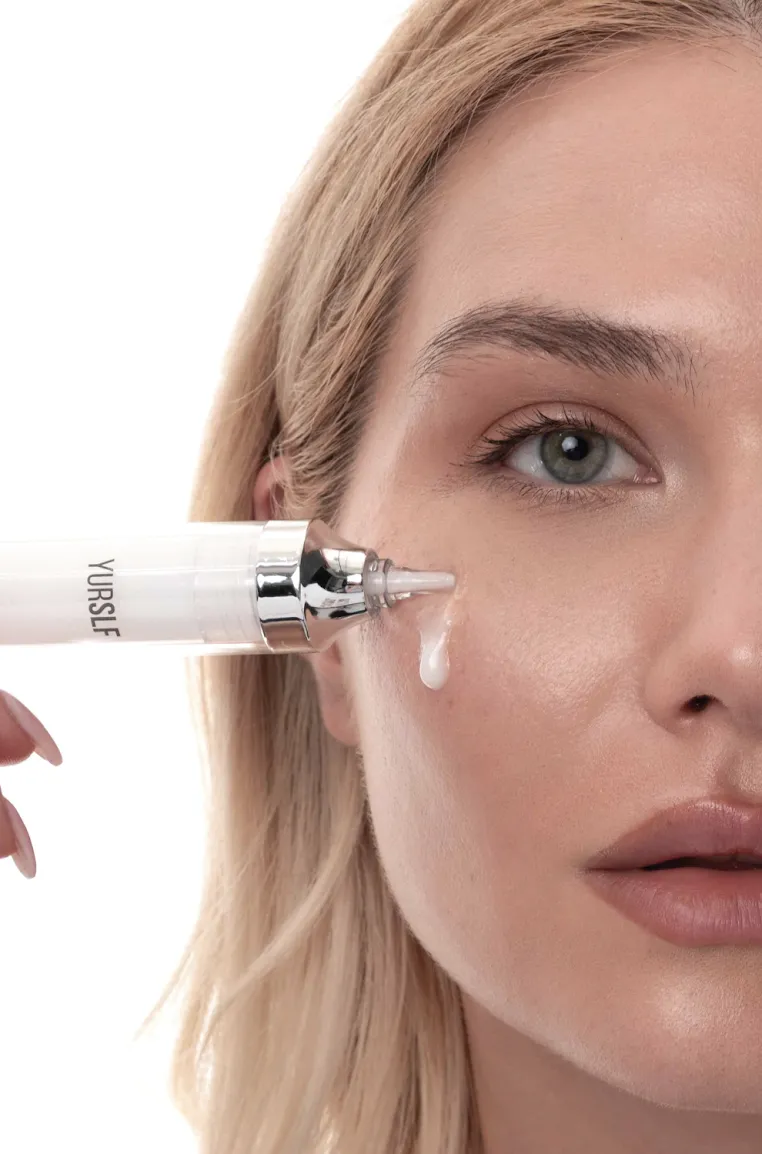 A close-up of a woman applying a white serum under her eye with a syringe-style applicator.