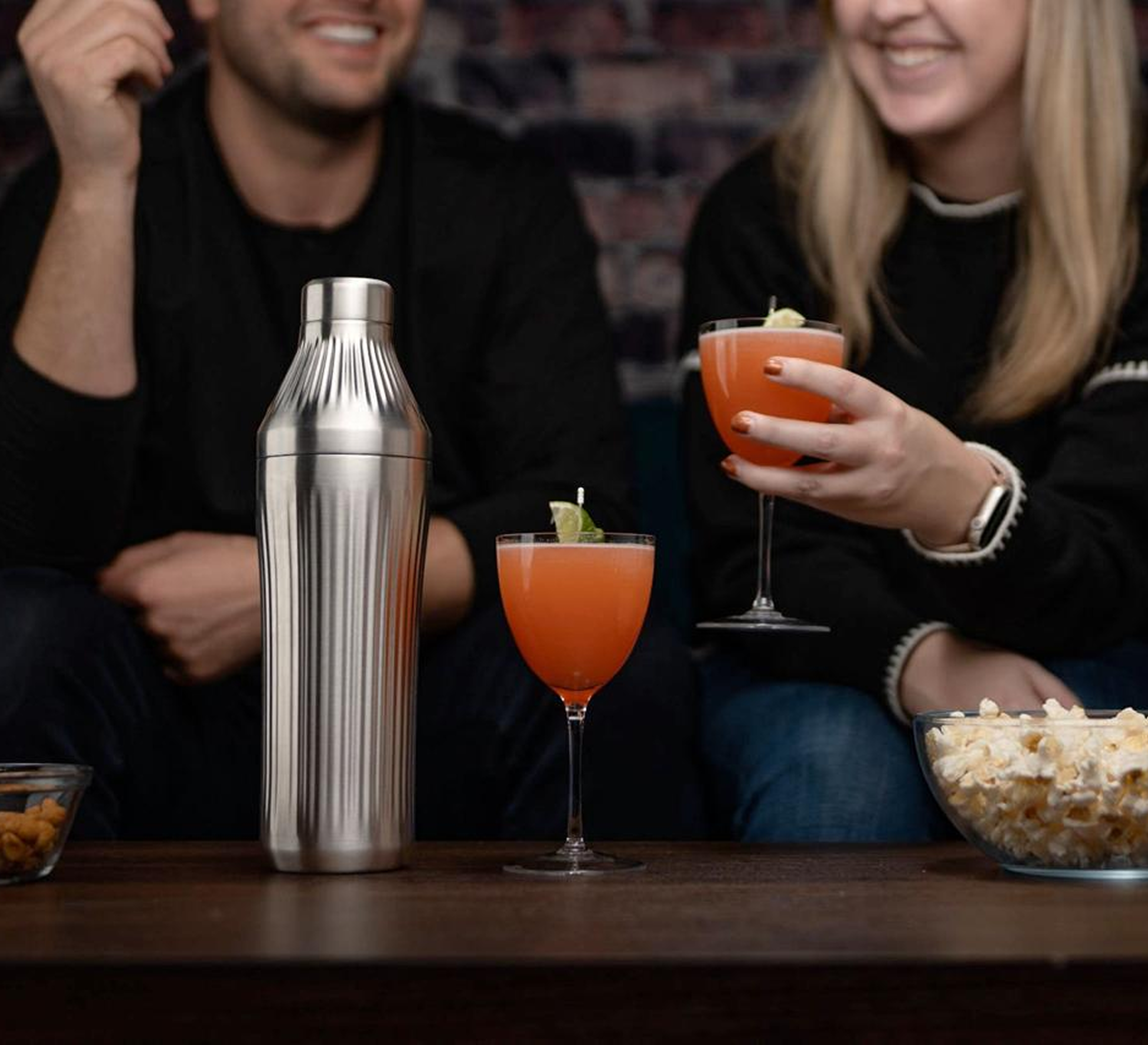 Two people with cocktails, a shaker, and snacks on a table.