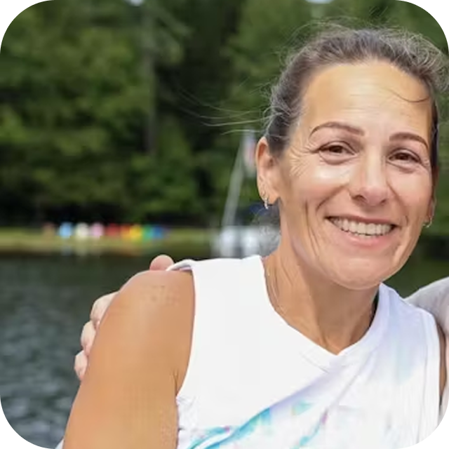 Smiling woman outdoors near a body of water and trees in the background.