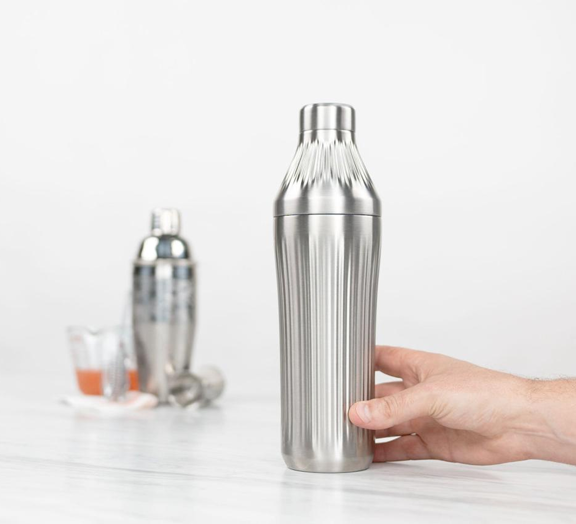 Hand holding a stainless steel cocktail shaker on a white surface.