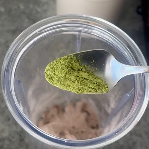 A spoon of green powder over a blender with brown powder inside.