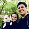 Two people smiling with a dog in a wooded area.