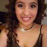 Smiling person with curly hair wearing a necklace and a patterned top.