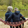 Two people sitting on a hammock by a lake, surrounded by trees.