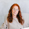 A person with long red hair and glasses sitting against a light background.