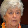 Elderly person with white hair and an orange scarf, speaking.