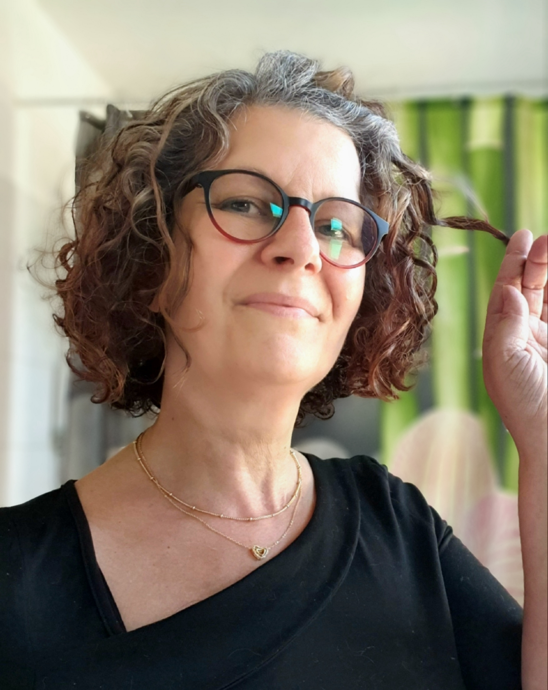 Person with curly hair and glasses, holding a strand, wearing a black top and gold necklace.
