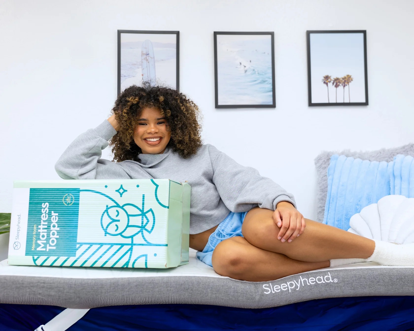 A smiling woman lies on a bed next to a Sleepyhead mattress topper box.