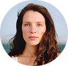 A circular headshot of a woman with long brown hair and light-colored eyes, with the sea in the background.