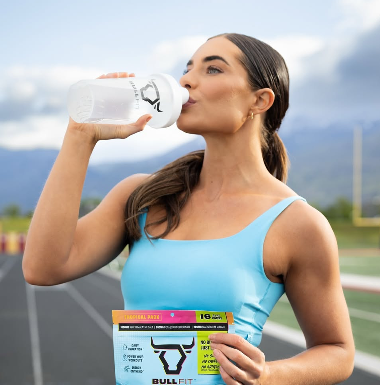 Woman in blue top drinking from a bottle on a track field.