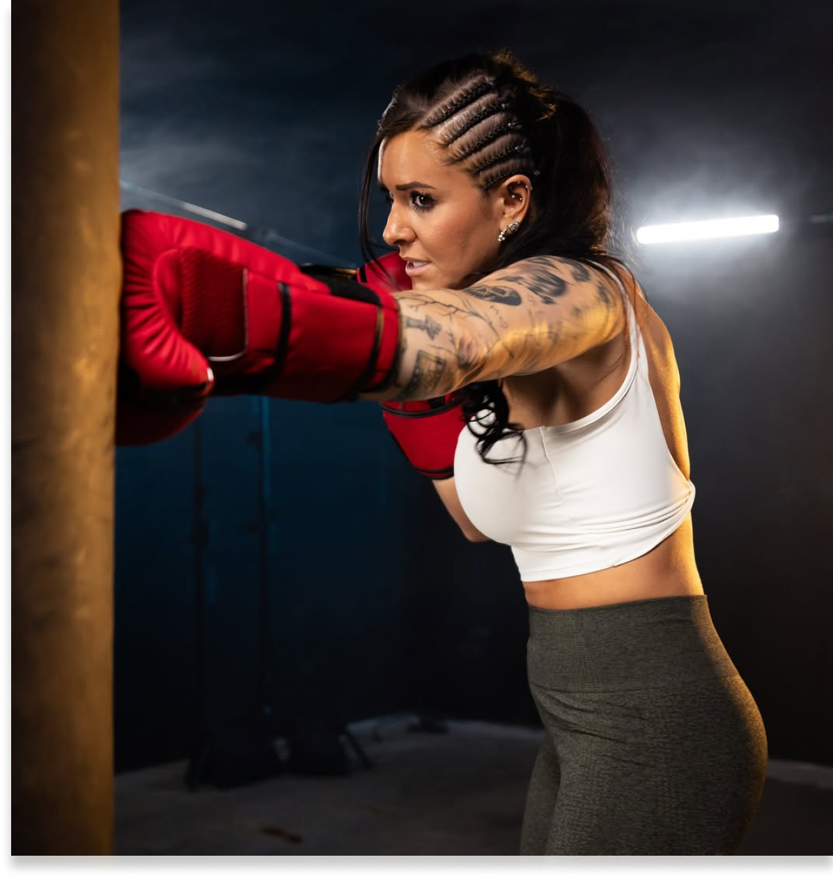 Woman with braided hair boxing in a gym, wearing red gloves and a white top.