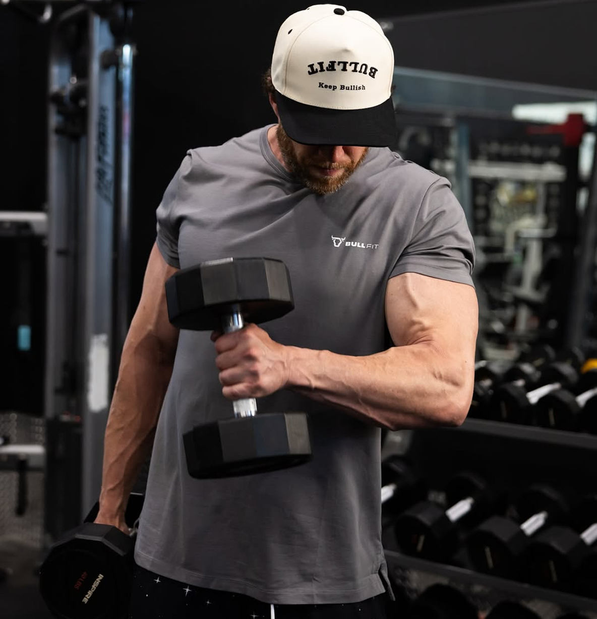 Man lifting dumbbells in a gym, wearing a cap and gray shirt.