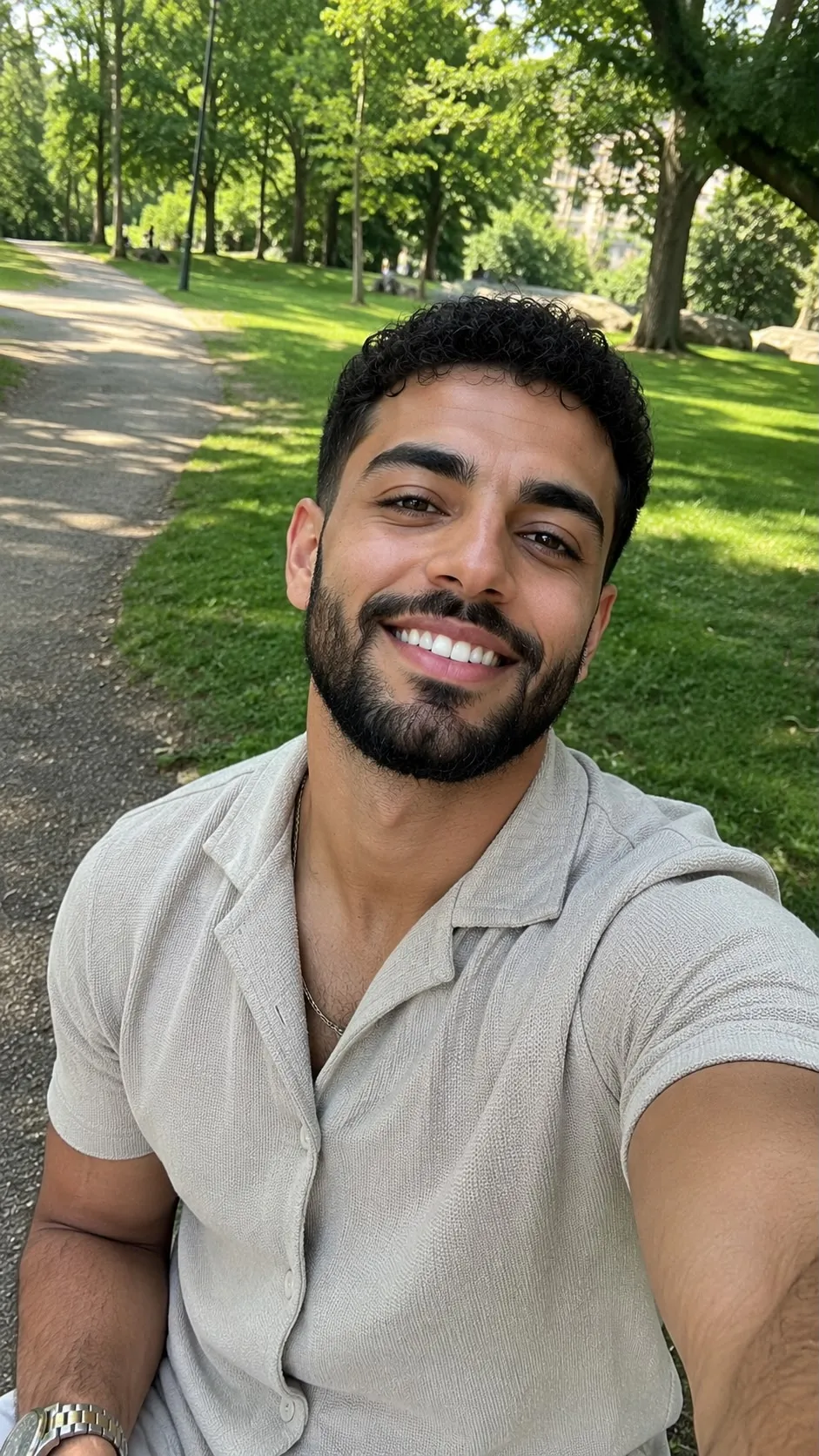 A smiling man with a beard takes a selfie while sitting in a park on a sunny day.