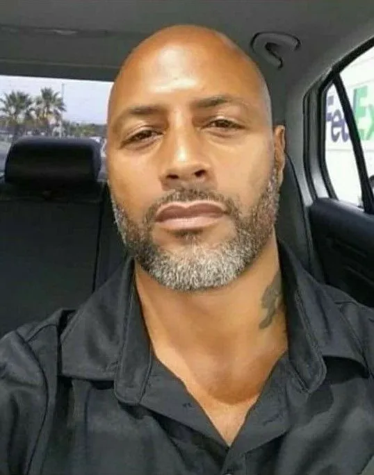 A close-up photo of a bald man with a salt-and-pepper beard wearing a black collared shirt inside a car.