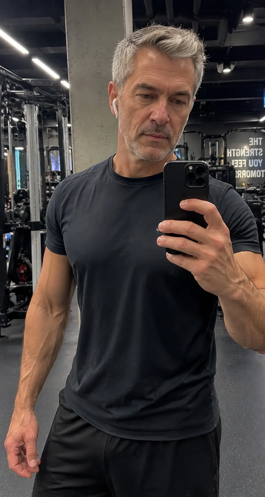 A fit, grey-haired man in a black t-shirt takes a mirror selfie in a gym.