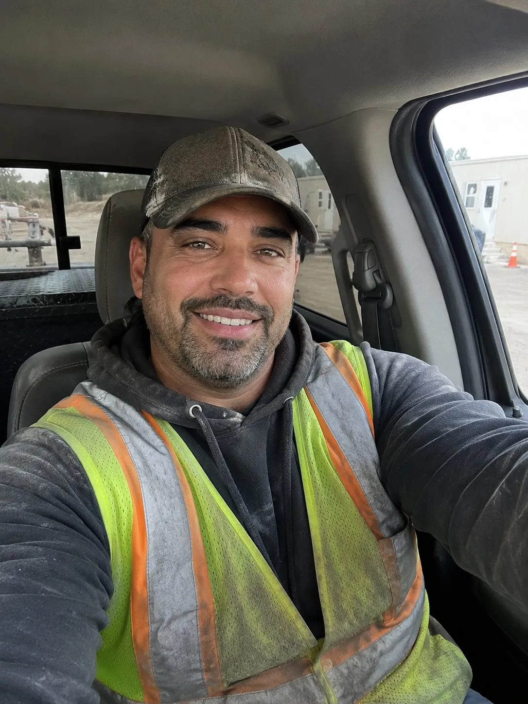 A smiling man in a high-visibility vest and a baseball cap takes a selfie in a vehicle.