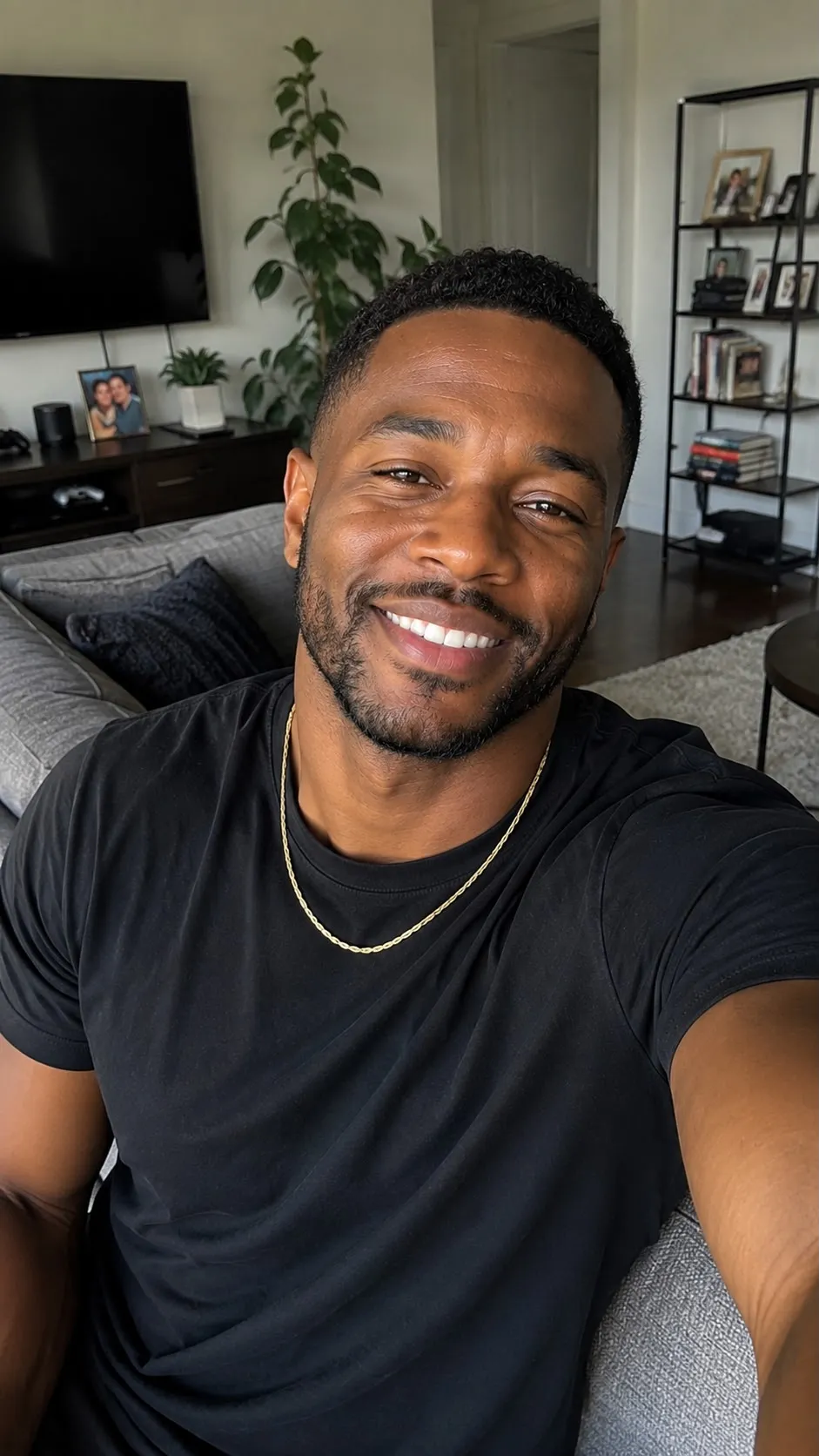 A smiling man in a black t-shirt and a gold chain takes a selfie in a living room.