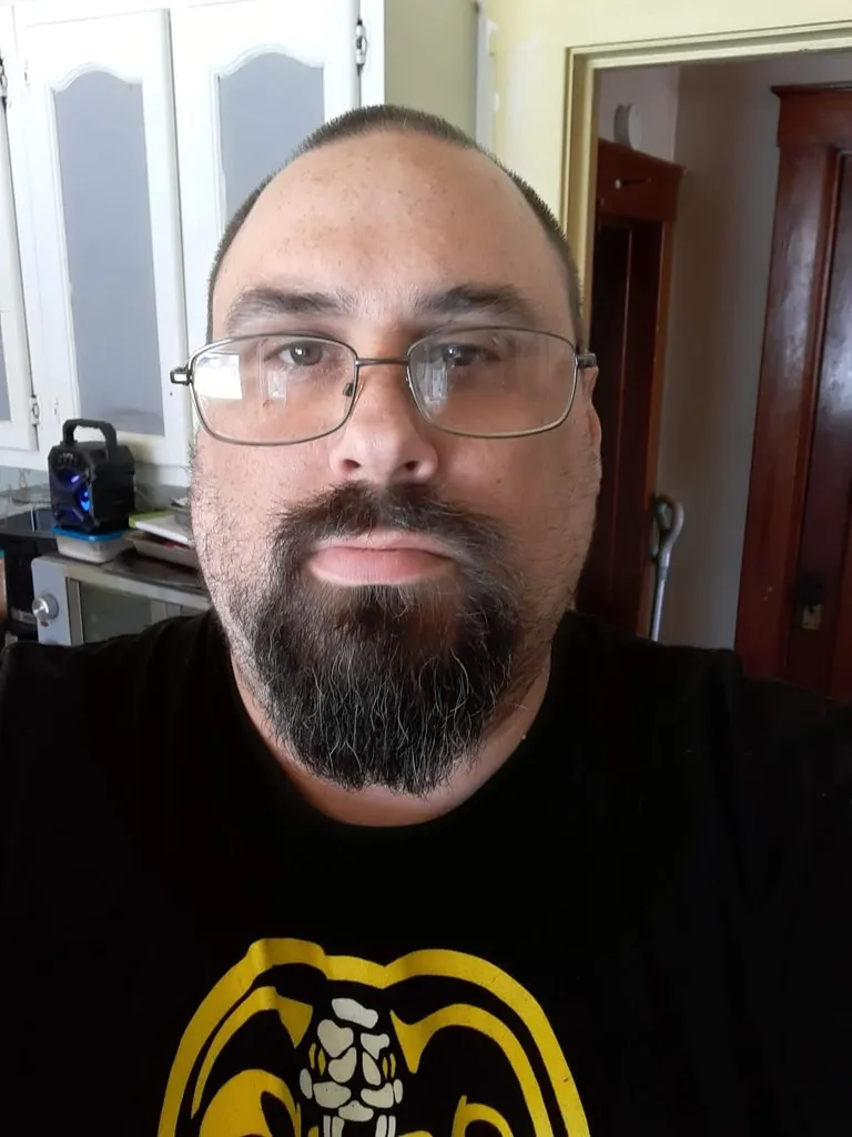 A close-up selfie of a man with glasses and a goatee wearing a black Cobra Kai t-shirt.