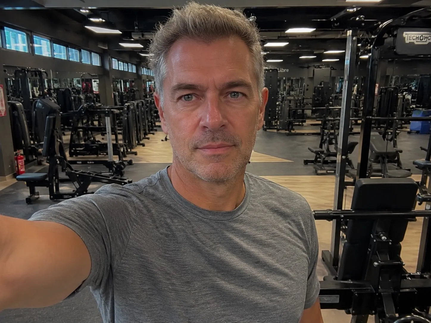 A man with graying hair and blue eyes takes a selfie in a gym.