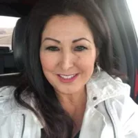 A woman with dark hair and a white jacket smiles while sitting in a car.