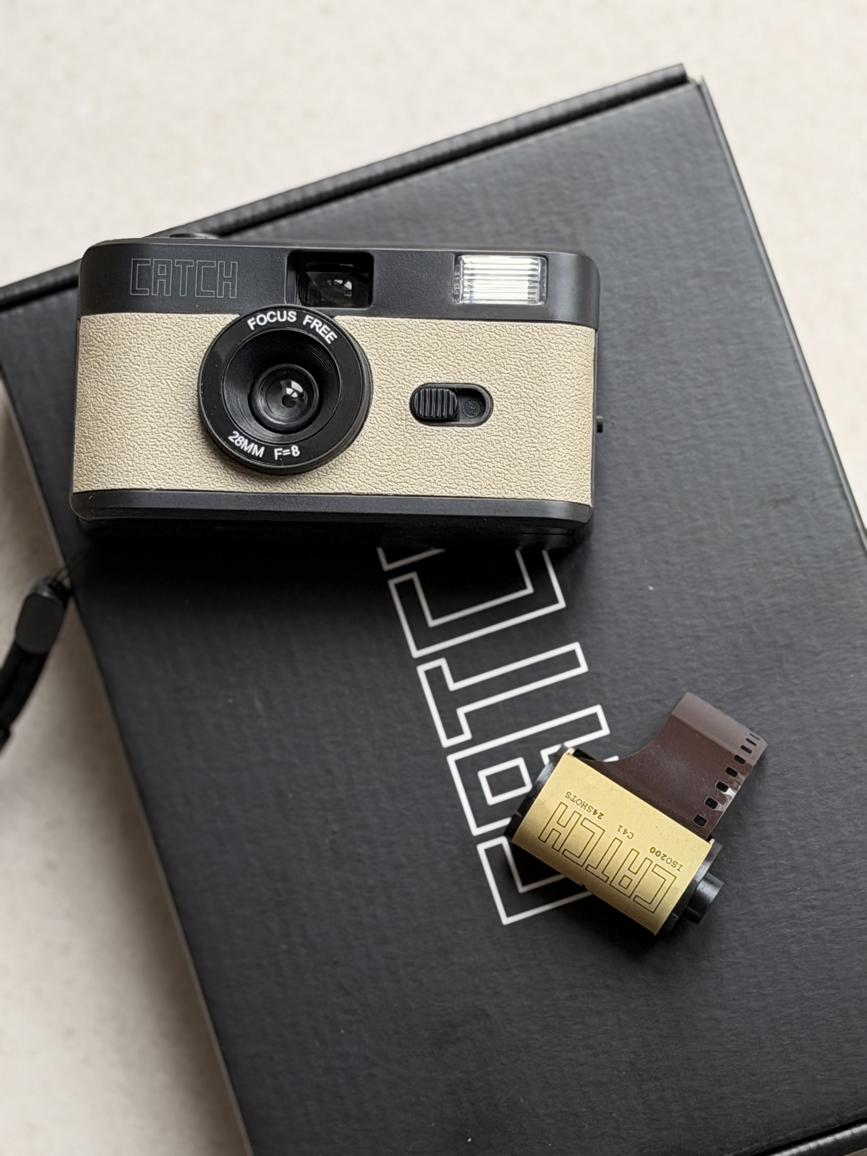 A beige and black Catch brand point-and-shoot camera sits on a black box next to a roll of film.