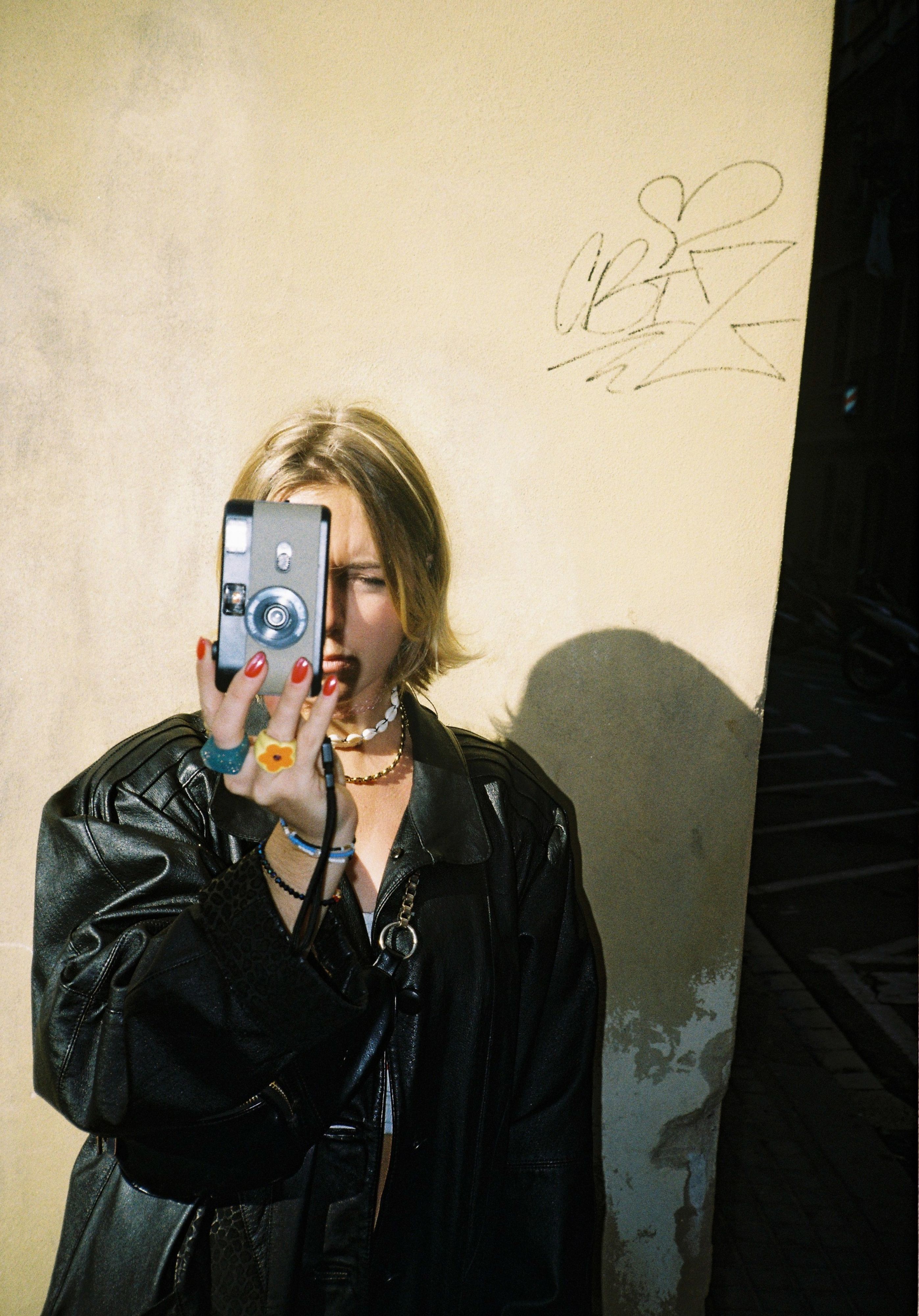 Person in leather jacket holding a camera, graffiti on wall.