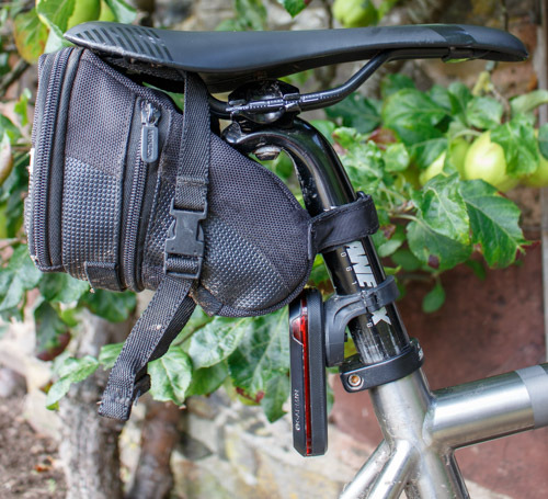 Bicycle seat with attached black storage bag and LED light, against a leafy background.