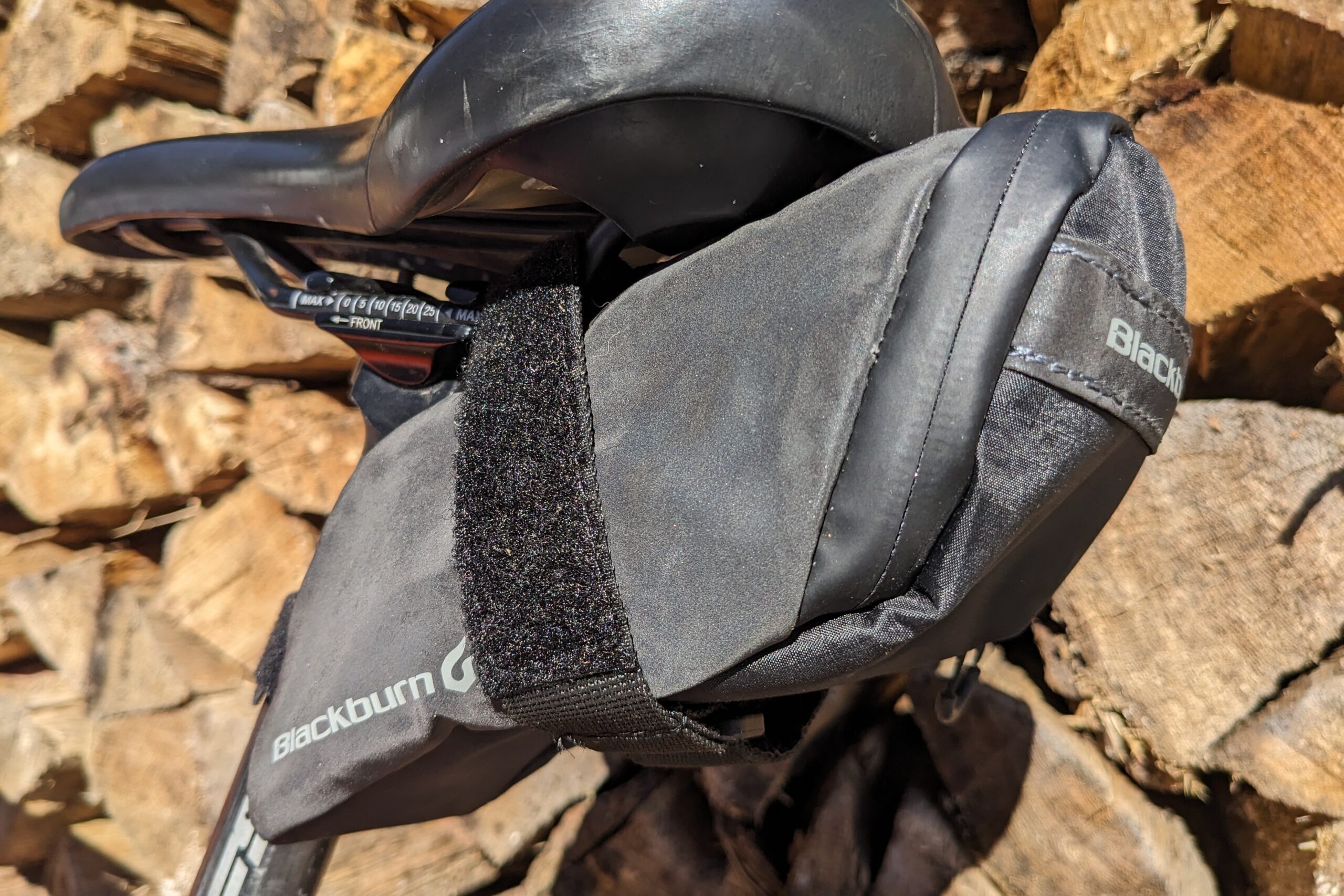 Close-up of a bicycle saddle with a black seat bag attached, against a wood pile background.