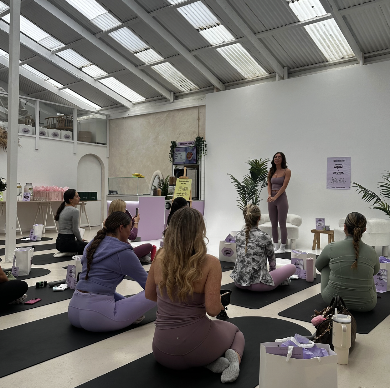 A woman in athletic wear addresses a group of women sitting on yoga mats in a bright studio.