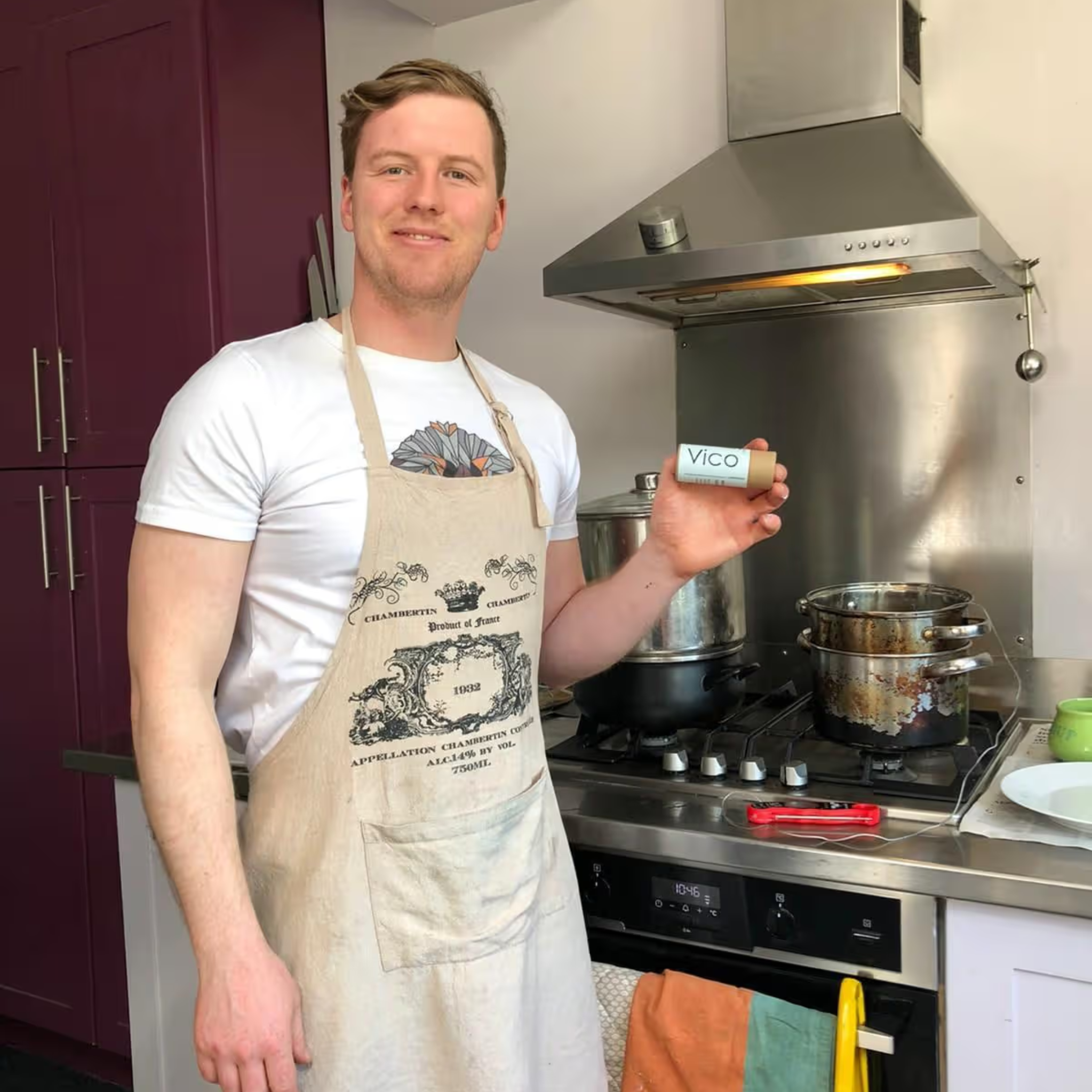 A man in an apron stands in a kitchen, smiling and holding a small product.