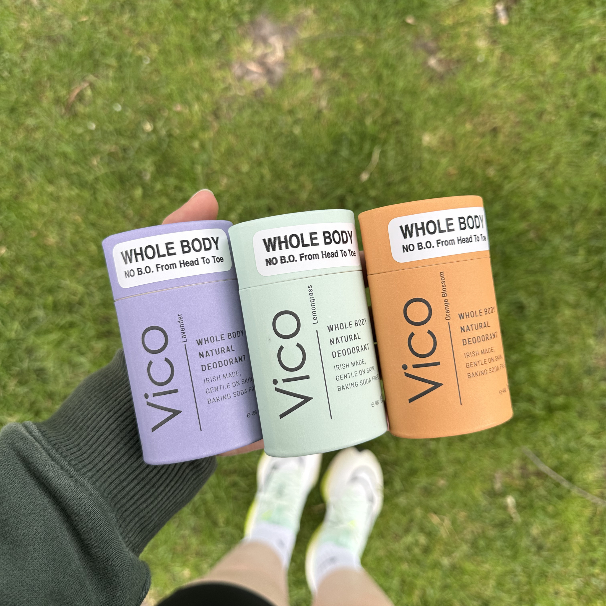 A hand holding three different colored tubes of Vico brand Whole Body deodorant over a grassy background.