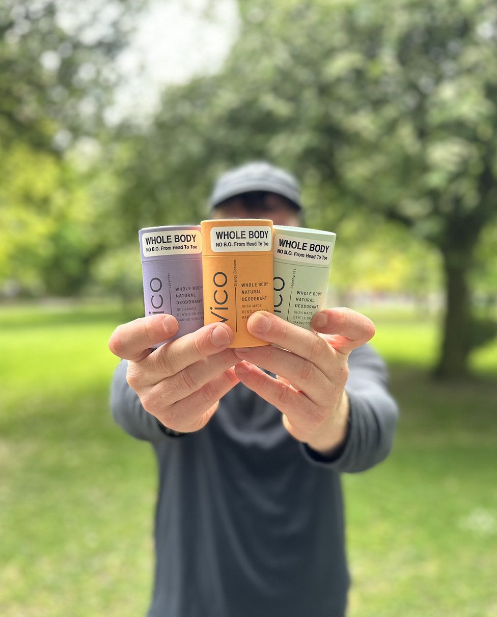 A person holds up three colorful tubes of Vico whole body deodorant in a park.