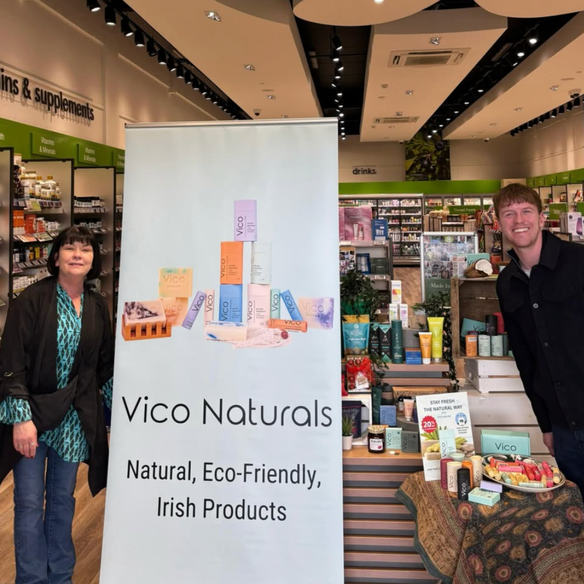 A woman and a man stand on either side of a Vico Naturals promotional banner inside a store.