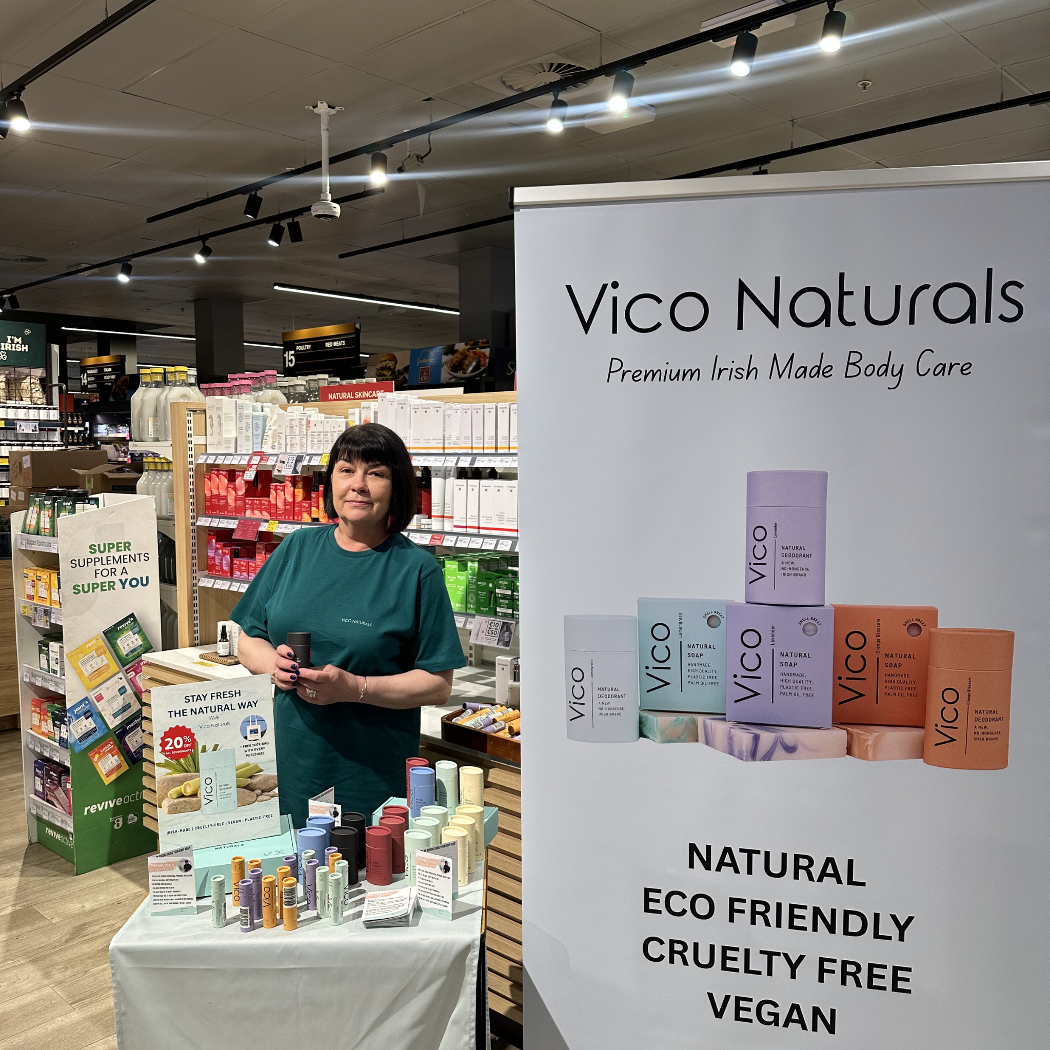 A woman stands behind a Vico Naturals promotional display stand inside a store.